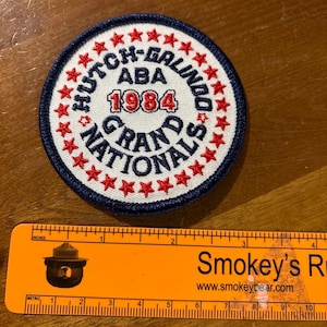 May include: A white and blue circular patch with red stars around the border. The text on the patch reads "Hutch-Galindo ABA 1984 Grand Nationals".
