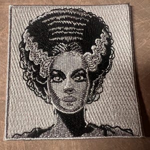 May include: A black and white embroidered patch featuring a close-up of the face of a woman with a hairstyle reminiscent of Frankenstein's monster. The patch is on a silver background.