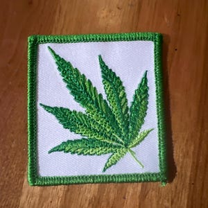 Puede incluir: Parche bordado verde con una hoja de marihuana sobre un fondo blanco y un borde verde.