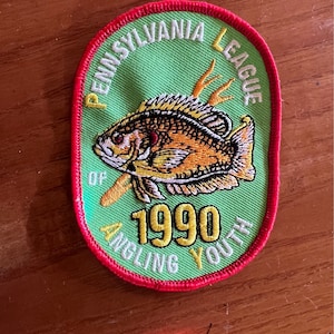 Pode incluir: Um remendo oval verde e vermelho com o texto "Pennsylvania League of Angling Youth" e o ano "1990". Um peixe amarelo e laranja com uma vara de pesca está no centro.