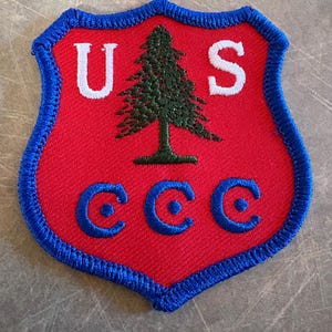 May include: A red shield-shaped patch with a blue border. The patch features the white letters "U" and "S" at the top, and "CCC" in blue at the bottom. A green tree is in the center.