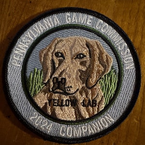 Puede incluir: Parche bordado circular con un Labrador Retriever amarillo. El texto "PENNSYLVANIA GAME COMMISSION" rodea la imagen, con "YELLOW LAB" y "2024 COMPANION" en el centro. El perro está sobre un fondo azul claro con hierba verde.