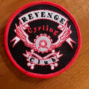 May include: A black and red embroidered patch with the words "Revenge Cycling Club" in white lettering. The patch features a pair of wings with a gear in the centre.