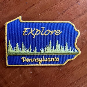 May include: An embroidered patch shaped like the state of Pennsylvania, featuring a blue background with yellow stitching. The patch reads "Explore Pennsylvania" and has a green line of trees across the middle.