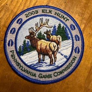 May include: A circular patch with a blue border and the text "2003 ELK HUNT" and "PENNSYLVANIA GAME COMMISSION". The patch features two elk in a snowy landscape with evergreen trees.
