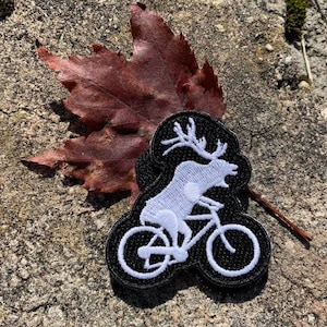 Könnte beinhalten: Weiß gestickter Aufnäher eines Rehs, das auf einem Fahrrad fährt, auf schwarzem Hintergrund.