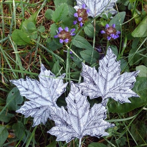 May include: Four white felt maple leaves with detailed stitching, arranged on a bed of green grass and purple wildflowers.