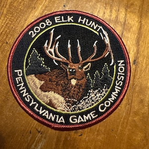 May include: Circular patch with a black background featuring a brown elk with large antlers, surrounded by text that reads "2008 ELK HUNT" and "PENNSYLVANIA GAME COMMISSION". The patch has a red border.