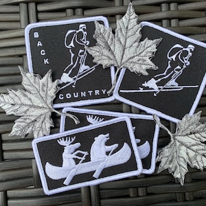 以下が含まれることがあります： 白い刺繍が施された黒い布のパッチが4つ。1つのパッチにはスキーヤーと「BACK COUNTRY」の文字が刺繍されている。2つのパッチにはスキーヤーが刺繍されている。1つのパッチには、カヌーに乗ったヘラジカとクマが刺繍されている。