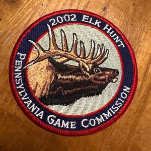 Puede incluir: Parche bordado circular con un diseño de alce. El parche presenta una ilustración detallada de un alce con grandes astas. El texto alrededor del borde dice "2002 ELK HUNT" y "PENNSYLVANIA GAME COMMISSION" en letras blancas sobre un fondo azul marino.