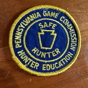 May include: Circular patch with a blue center and yellow border. The text "PENNSYLVANIA GAME COMMISSION" and "HUNTER EDUCATION" are in yellow. The center features a yellow keystone and the words "SAFE HUNTER".