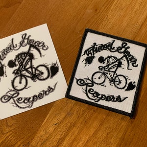 May include: A white sticker and a white patch with black embroidery, both featuring a skeleton riding a bicycle with a scythe. The text "Tired Year Reapers" is written in a stylized font around the image.