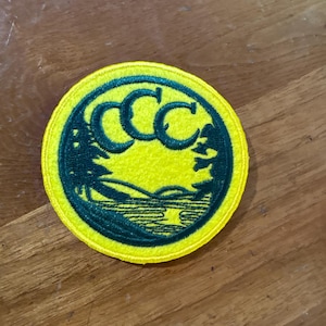 May include: A circular yellow patch with a green border and design. The design features three green "C" letters and a landscape scene with trees and water. The patch is likely an emblem or insignia.