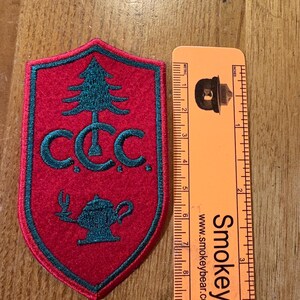 May include: A shield-shaped red patch with dark teal embroidery featuring a pine tree, the letters C.C.C., and an oil lamp. An orange ruler is placed next to the patch, showing it measures approximately 3.5 inches in total vertical height.
