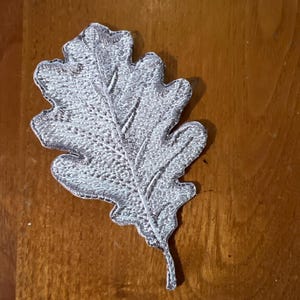May include: A silver embroidered oak leaf patch. The leaf is detailed with a textured, raised design.