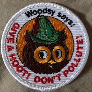 Puede incluir: Un parche bordado blanco con un borde rojo que presenta un búho de dibujos animados con un sombrero verde con una pluma roja. El búho tiene grandes ojos amarillos y un cuerpo marrón. El texto "Woodsy says: Give a hoot! Don't pollute!" está escrito en rojo alrededor del búho.