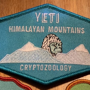 Op de afbeelding: Een turquoise blauwe geborduurde patch met witte stiksels die een Yeti in de Himalaya voorstelt. De tekst "YETI" staat bovenaan de patch, en "CRYPTOZOOLOGY" onderaan.