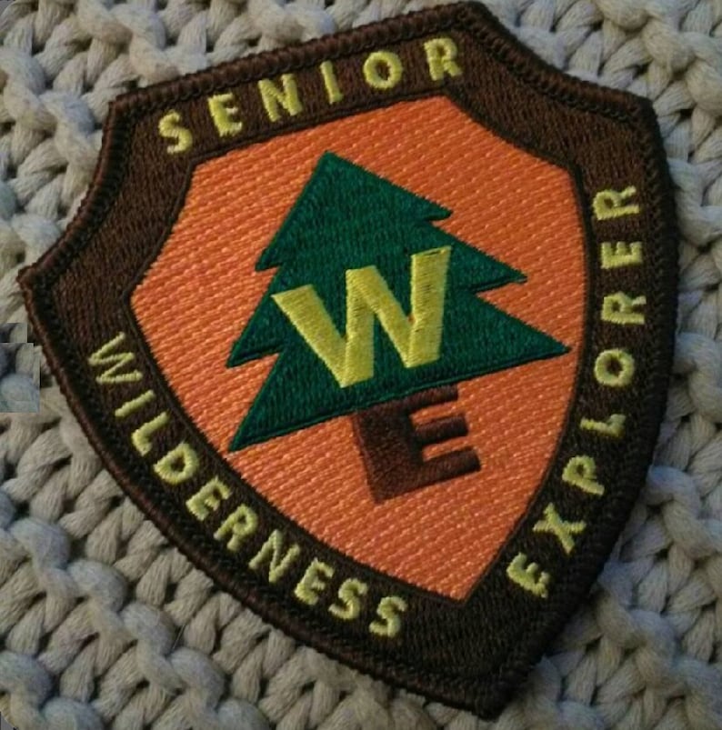 May include: A brown and orange embroidered patch with the text "Senior Wilderness Explorer" and a green pine tree with a yellow "W" inside.