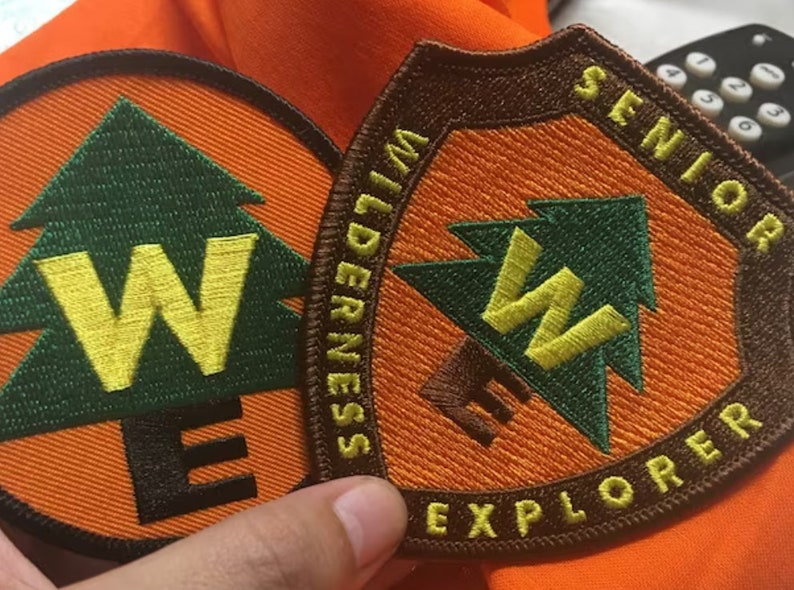 May include: Two Wilderness Explorer patches, one with the text "Senior Wilderness Explorer" and the other with the text "Wilderness Explorer". Both patches feature a green pine tree with a yellow "W" inside and an orange background.