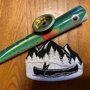 May include: A green and white fish-shaped pipe with a gold mouthpiece. The pipe is lying on a wooden surface next to a white and black embroidered patch with a mountain scene and a canoe.