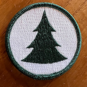 May include: A green embroidered pine tree patch on a white background with a green stitched border.