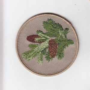 Puede incluir: Un parche redondo beige bordado con una rama de pino verde y dos piñas marrones.