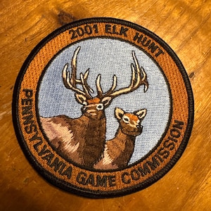 May include: A circular patch with a brown and tan border. The patch features two elk, a bull and a cow, in a blue circle. The text "2001 ELK HUNT" and "PENNSYLVANIA GAME COMMISSION" are printed on the patch.
