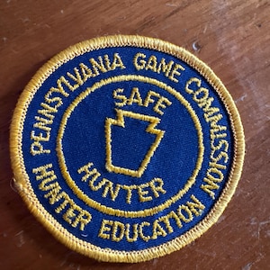 May include: A circular patch with a blue center and gold embroidered text and border. The text reads "PENNSYLVANIA GAME COMMISSION" around the top and right, and "HUNTER EDUCATION" around the bottom. A gold keystone shape is in the center, with the word "SAFE" above it.
