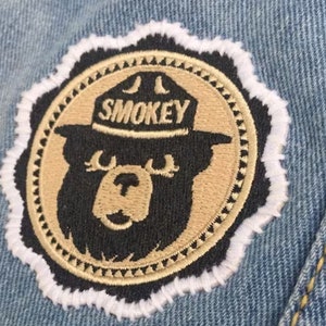 May include: A tan and black embroidered patch featuring Smokey Bear wearing a ranger hat with the text "SMOKEY" on it. The patch is surrounded by a white border and a tan scalloped border.