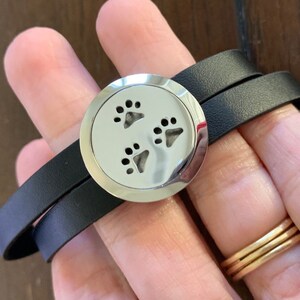 May include: A black leather wrap bracelet with a silver-colored, round charm. The charm features three paw prints. The bracelet wraps around the wrist twice.