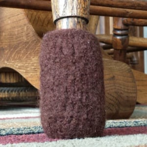 May include: A close-up of a brown chair leg with a dark brown, cylindrical furniture protector. The protector is made of a fuzzy material and is designed to prevent scratches on floors. The background shows other wooden chair parts.
