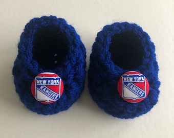 May include: A pair of blue crocheted baby booties with a red, white, and blue New York Rangers logo on each bootie.