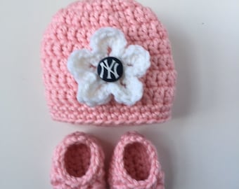 Puede incluir: Un gorro de bebé de ganchillo rosa con una flor de ganchillo blanca y un botón blanco y negro con las letras "NY". El conjunto también incluye dos botines de ganchillo rosa.
