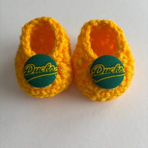 May include: A pair of yellow crocheted baby booties with green buttons that say "Ducks" in yellow lettering.