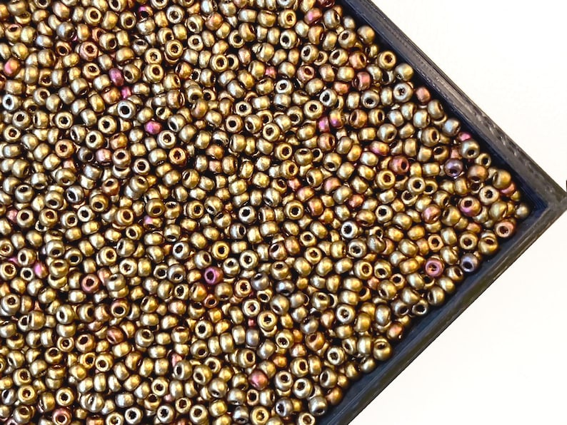 Puede incluir: Una imagen de primer plano de un contenedor lleno de peque&ntilde;as cuentas redondas de color dorado. Las cuentas tienen un brillo met&aacute;lico y una ligera calidad iridiscente.