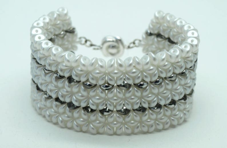 May include: A white beaded bracelet with a silver clasp. The bracelet is made of small, round beads that are arranged in a pattern. The bracelet is a wide style and has a silver clasp.