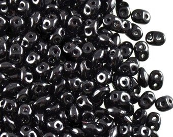 Può includere: Una collezione di perline nere lucide a due fori, ognuna caratterizzata da una forma allungata e arrotondata con due fori circolari distinti posizionati fianco a fianco. Le perline hanno una superficie liscia e riflettente che cattura la luce, creando riflessi bianchi brillanti sul colore scuro e uniforme del materiale in vetro.