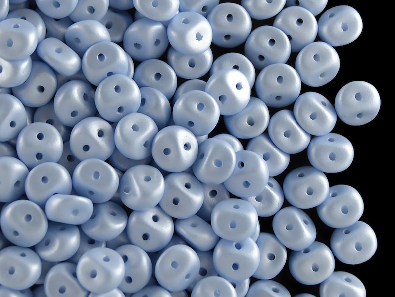 Pu&ograve; includere: Un mucchio di perline di vetro blu chiaro con due fori in ogni perlina. Le perline sono lisce e hanno una finitura leggermente iridescente.