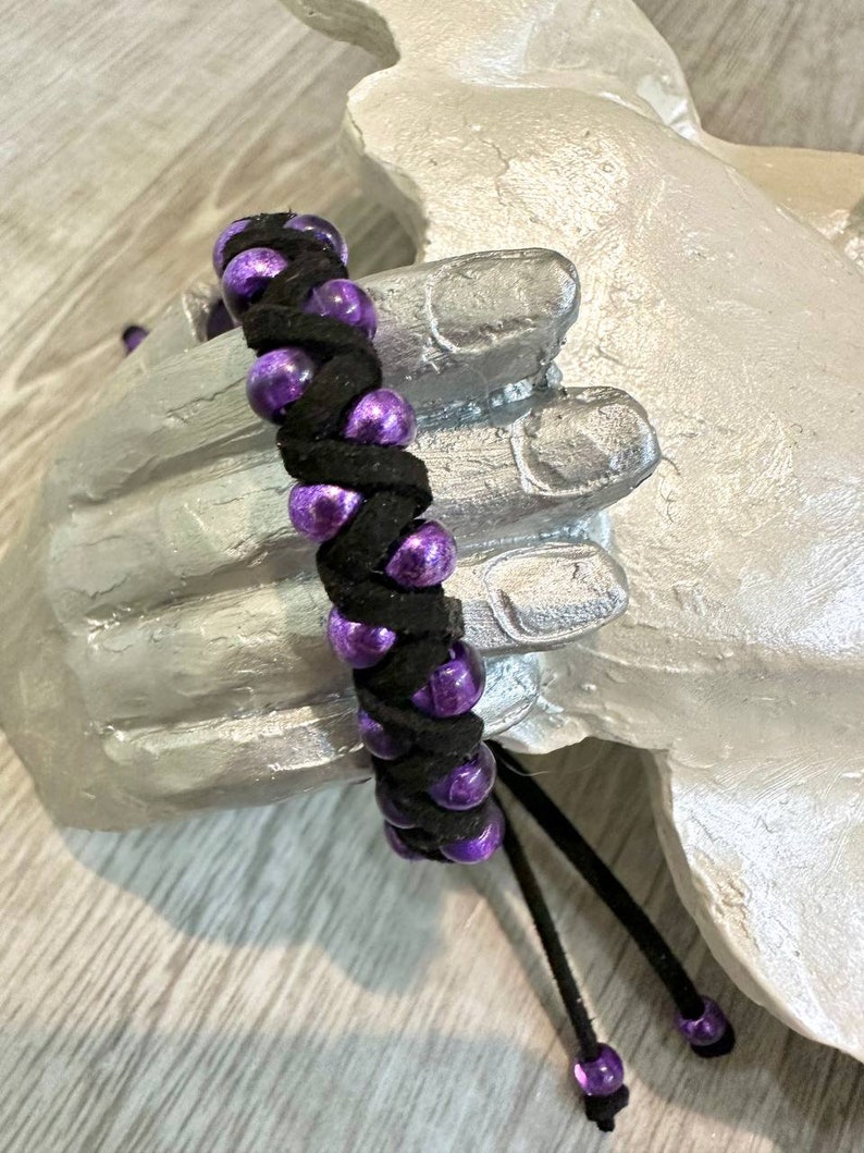 Op de afbeelding: Een zwarte gevlochten armband met paarse kralen. De armband heeft een kwast aan het einde.