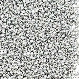 May include: A large collection of small, round, metallic silver seed beads. Each bead features a smooth, reflective surface and a central hole, creating a uniform appearance. The beads are densely packed together, showing a consistent colour.