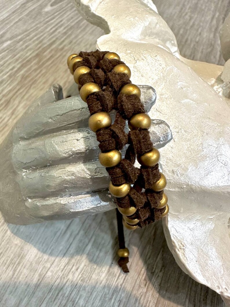 May include: A brown leather bracelet with gold beads woven into the design. The bracelet has a simple tie closure.