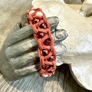Op de afbeelding: Een gevlochten leren armband met roze en bruine kralen. De armband is geweven met een decoratief patroon.