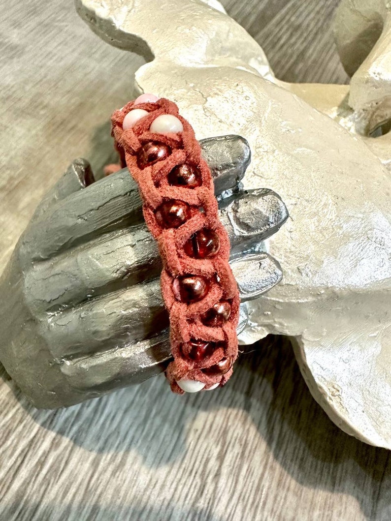 Op de afbeelding: Een gevlochten leren armband met roze en bruine kralen. De armband is geweven met een decoratief patroon.