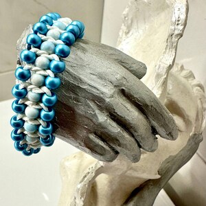 May include: A blue and white beaded bracelet with a woven design. The bracelet is made of small, round beads and is displayed on a white hand sculpture.