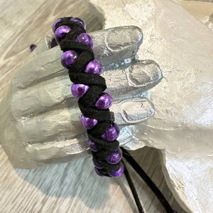 May include: A black braided bracelet with purple beads. The bracelet has a tassel on the end.
