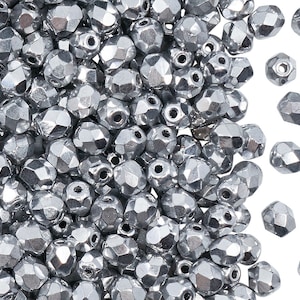 May include: A large collection of small, metallic silver-coloured beads with a faceted, multi-sided surface. Each bead features a central hole for threading, and the overall appearance is reflective and uniform, showcasing a dense pile of loose components.