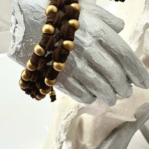Op de afbeelding: Een bruin leren armband met gouden kralen die in het ontwerp zijn geweven. De armband is vastgebonden met een bruin leren koord.
