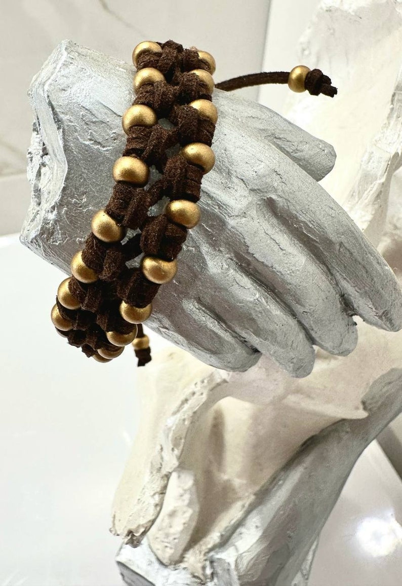 Op de afbeelding: Een bruin leren armband met gouden kralen die in het ontwerp zijn geweven. De armband is vastgebonden met een bruin leren koord.