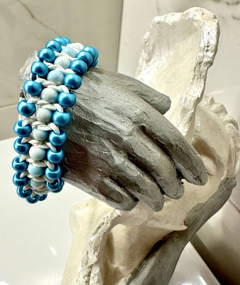 Op de afbeelding: Een blauwe en witte kralen armband met een geweven ontwerp. De armband is gemaakt van kleine, ronde kralen en wordt getoond op een wit handbeeld.