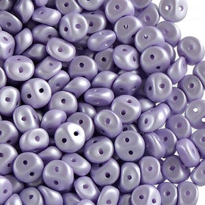 May include: A pile of small, light purple, two-hole beads. The beads are smooth and have a pearlescent finish.